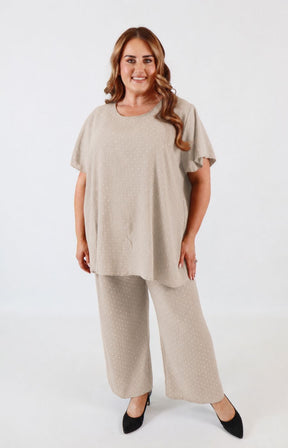 A woman modeling a matching beige knit lounge set consisting of a short-sleeve crewneck top and wide-leg pants, paired with black pointed-toe heels against a plain white studio background.