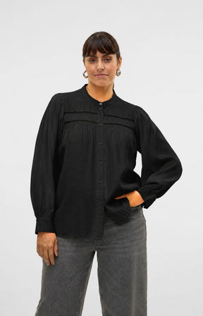 A woman wearing a black long-sleeve button-up blouse with pleated detailing across the chest and gray trousers, standing against a light gray studio background.