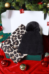 Black knit beanie with pom-pom and leopard print scarf on a red fabric background with Christmas decorations.