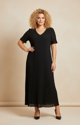 A full-length front view of a woman with shoulder-length curly blonde hair wearing a black pleated midi dress with short flutter sleeves. She stands in black high heels against a soft beige studio background with warm, even lighting.