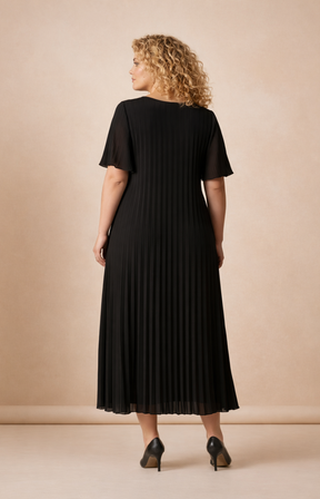 A full-length rear view of a woman with shoulder-length curly blonde hair wearing a black pleated midi dress with short flutter sleeves. She stands in black high heels against a soft beige studio background with warm, even lighting.