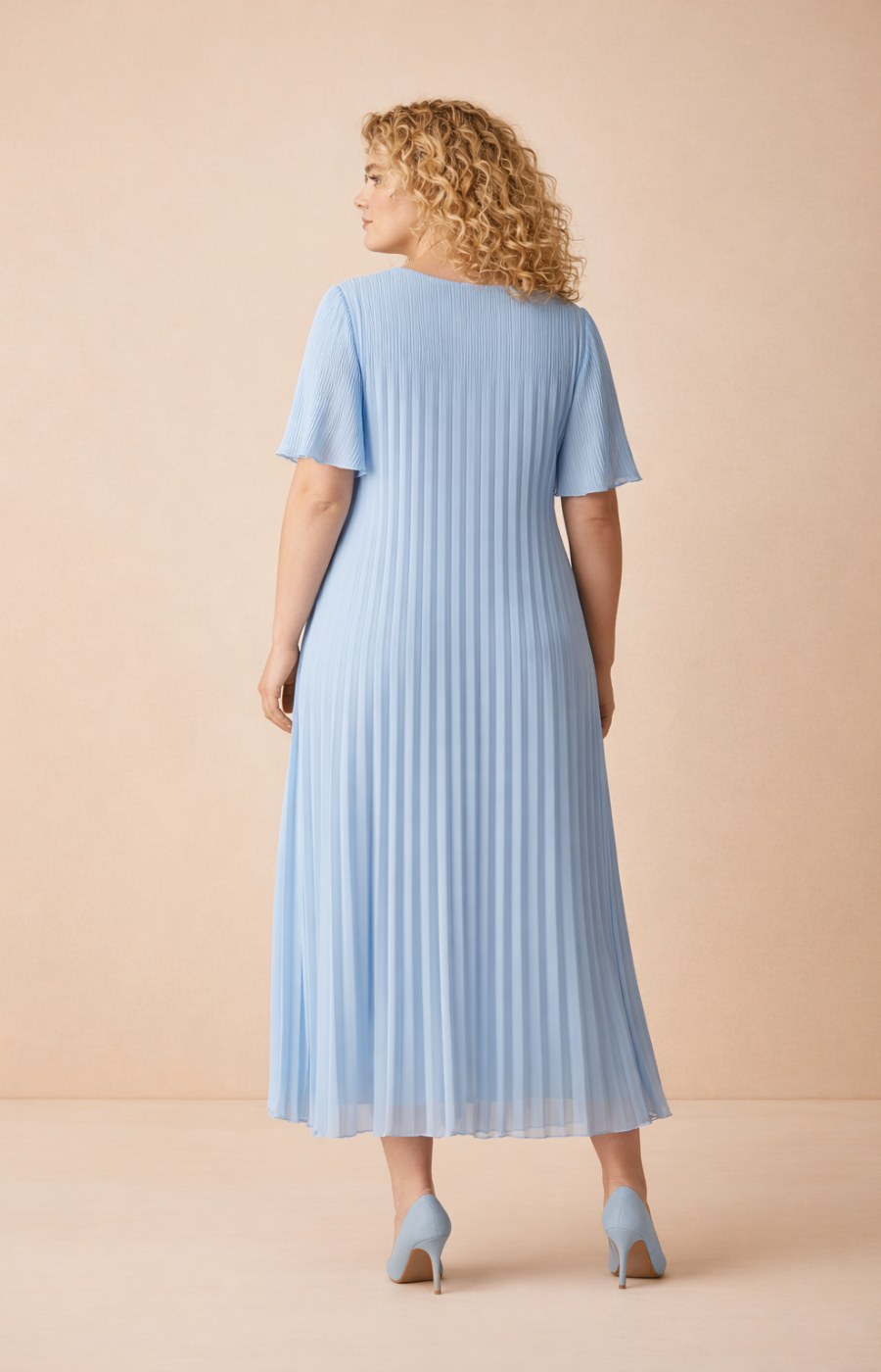 A full-length rear view of a woman with shoulder-length curly blonde hair wearing a blue pleated midi dress with short flutter sleeves. She stands in blue high heels against a soft beige studio background with warm, even lighting.