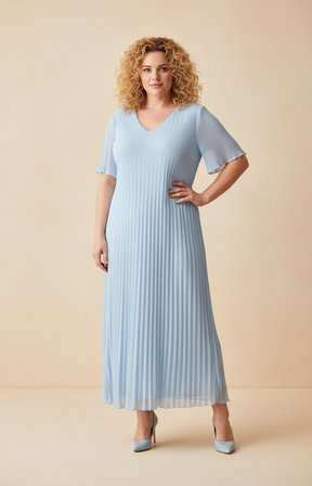 A full-length front view of a woman with shoulder-length curly blonde hair wearing a blue pleated midi dress with short flutter sleeves. She stands in blue high heels against a soft beige studio background with warm, even lighting.