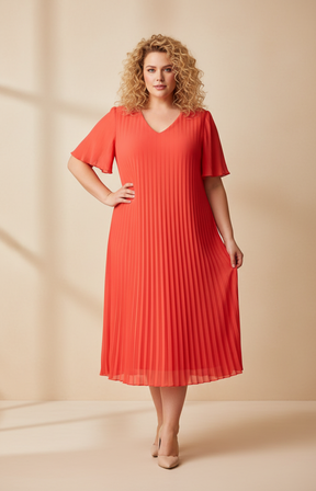 A full-length front view of a woman with shoulder-length curly blonde hair wearing a coral pleated midi dress with short flutter sleeves. She stands in high heels against a soft beige studio background with warm, even lighting.