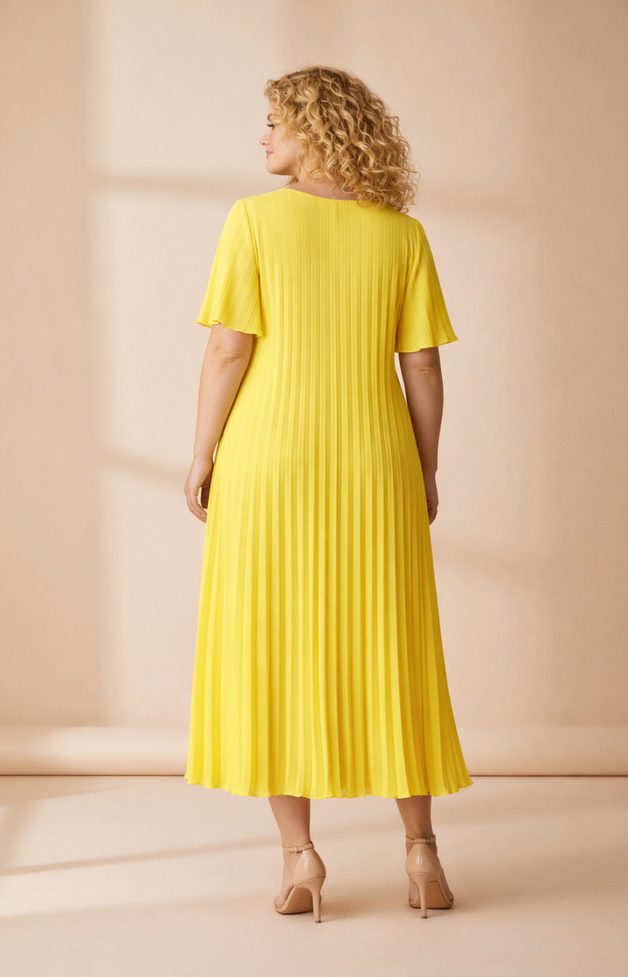 A full-length rear view of a woman with shoulder-length curly blonde hair wearing a yellow pleated midi dress with short flutter sleeves. She stands in cream high heels against a soft beige studio background with warm, even lighting.