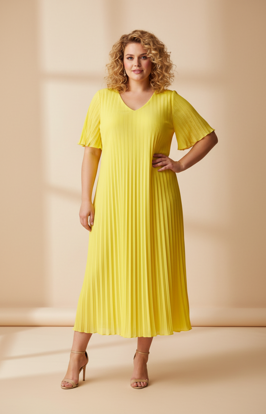 A full-length front view of a woman with shoulder-length curly blonde hair wearing a yellow pleated midi dress with short flutter sleeves. She stands in cream high heels against a soft beige studio background with warm, even lighting.