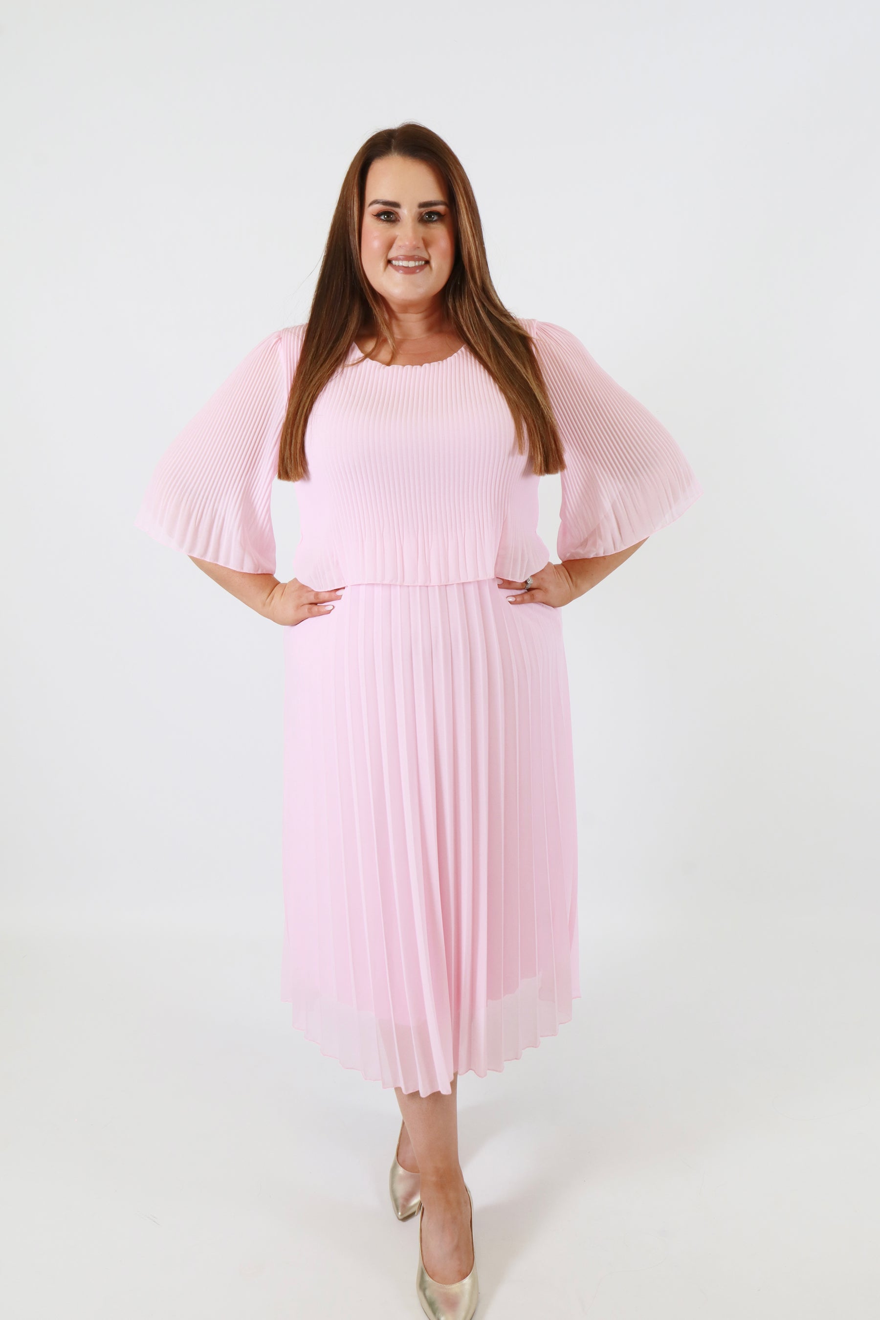 Plus size woman wearing a pink pleated dress with gold shoes on a white background