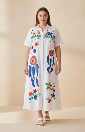 Woman wearing the Fern Floral Dress in White, a white dress with colorful floral and abstract designs on a beige background