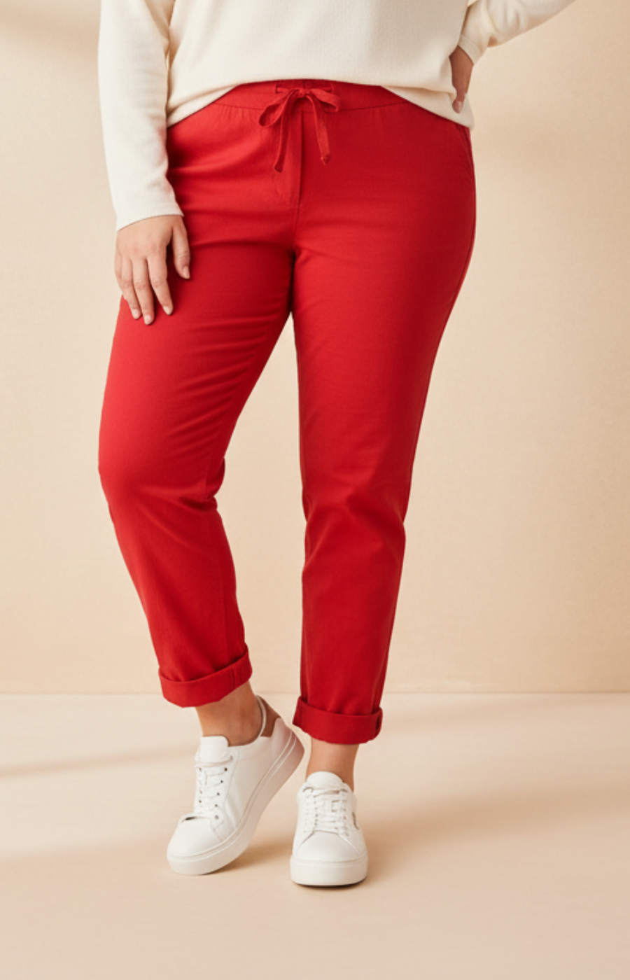 A plus size woman wearing red joggers and a cream long sleeve top on a beige background