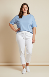 A plus size woman wearing white trousers, blue t-shirt and white trainers on a beige background