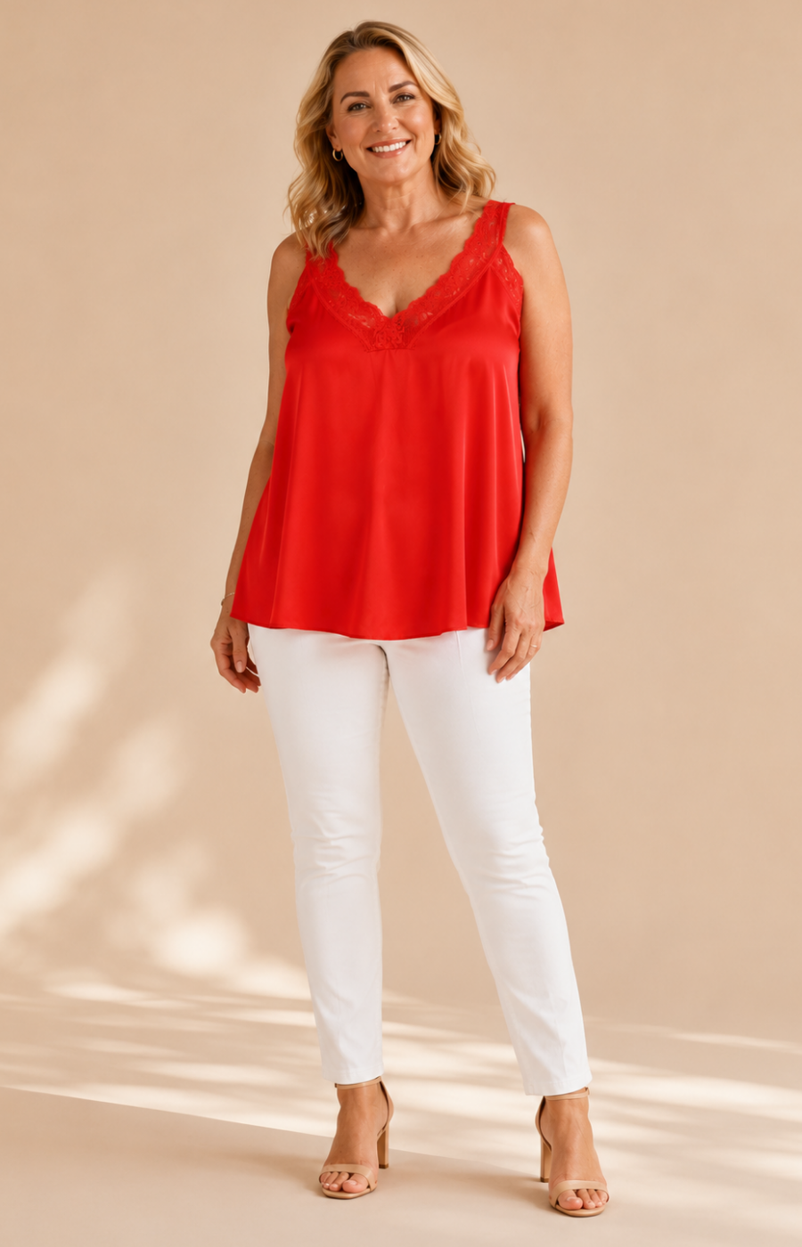 A woman modeling a bright red sleeveless camisole top with a V-neck and scalloped lace trim, paired with white pants and beige heels, against a warm beige studio backdrop.