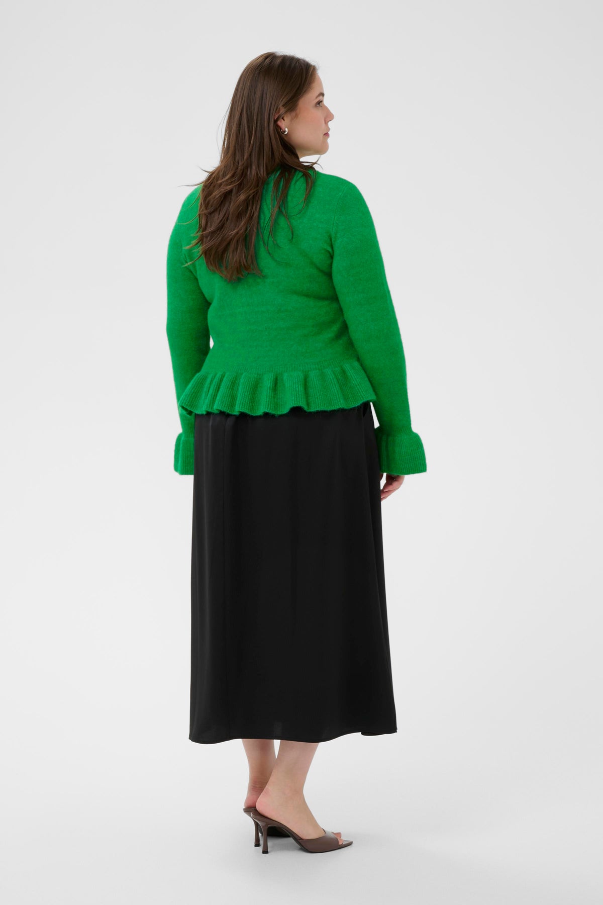 Woman to the back wearing a green cardigan with long sleeves and ruffle detailing on the hem and cuffs and black skirt on a white background