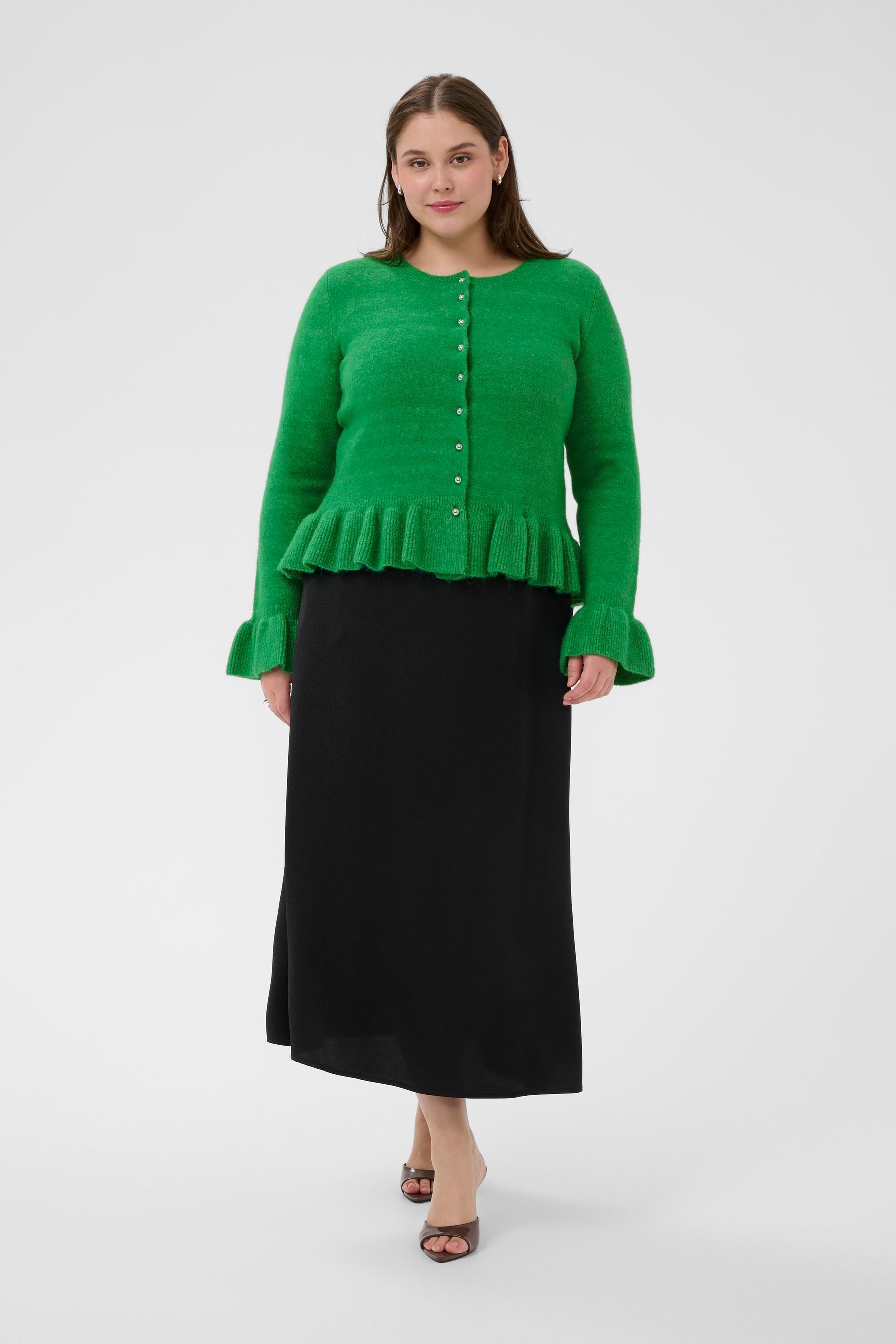 Woman wearing a green cardigan with a round neckline, silver buttons and a ruffle hem on the edge and on the cuffs and black skirt on a white background