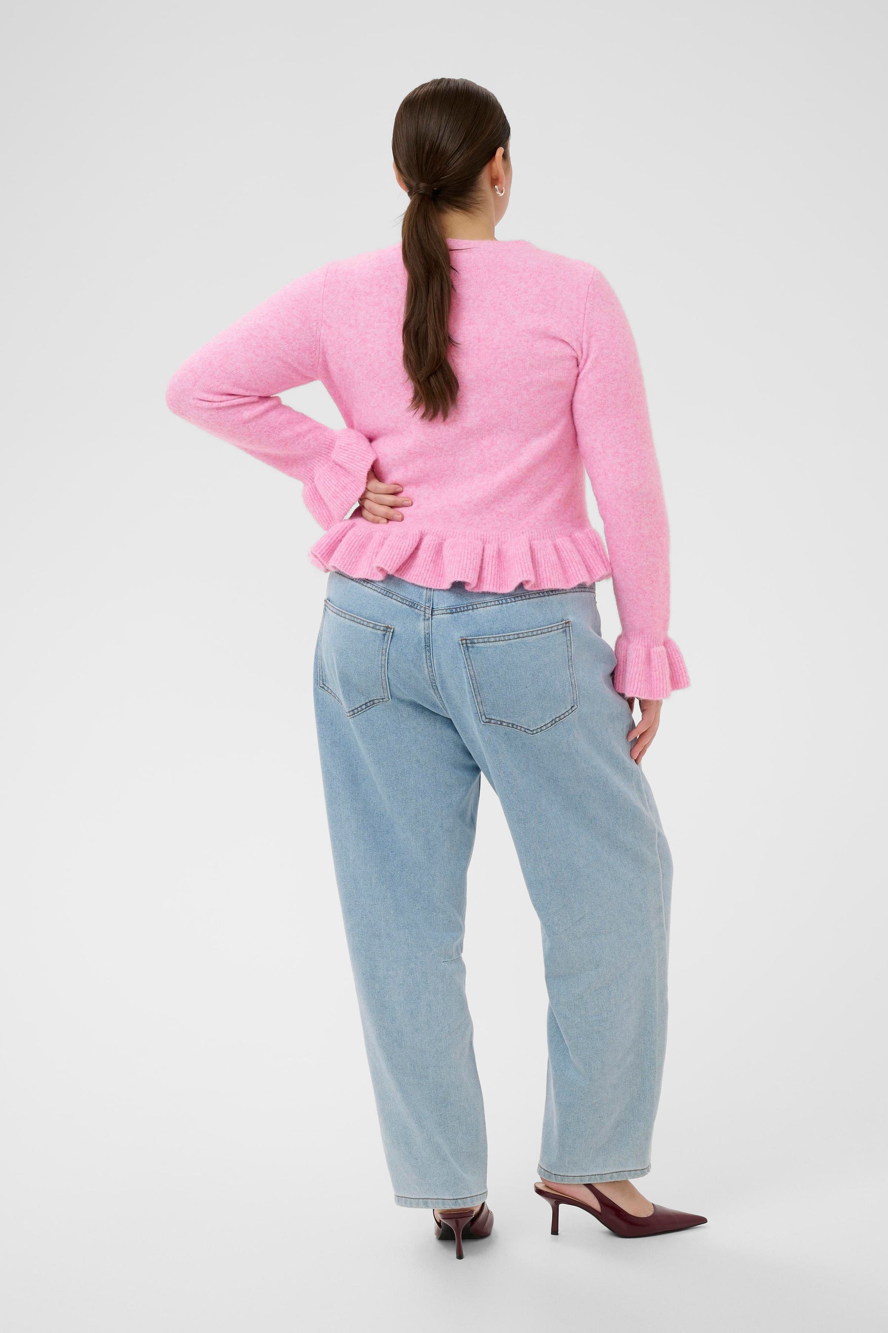 Person wearing a pink cardigan with long sleeves with ruffle detailing on the cuffs and hem and light blue jeans on a white background