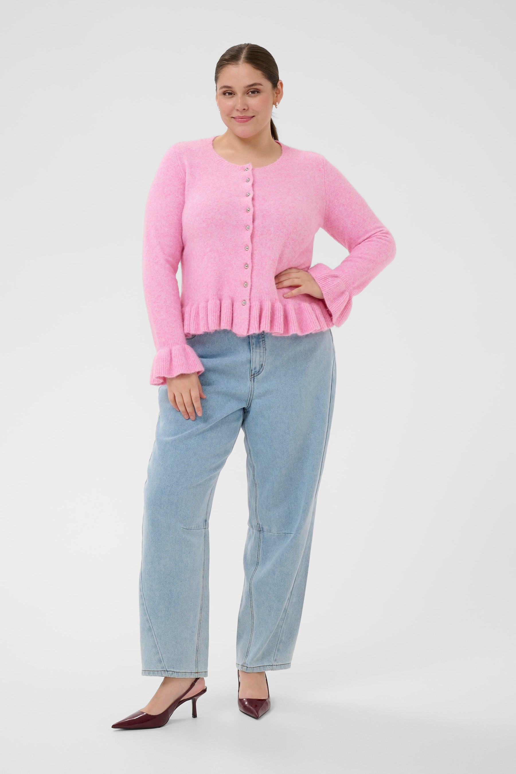 Woman wearing a pink cardigan with a round neckline and long sleeves, silver buttons and ruffle detailing on the hem and cuffs and light blue jeans on a white background