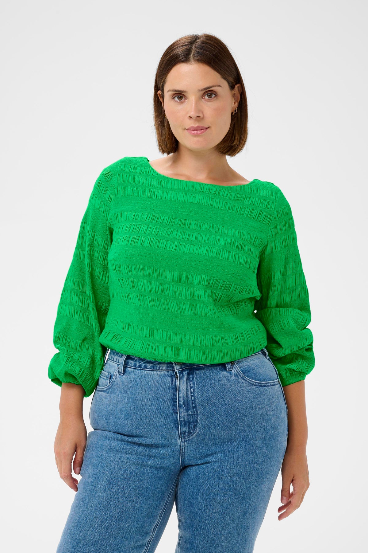 Woman wearing the Kaffe Curve Jourtney Blouse in Green, a green textured blouse with long sleeves and a boat neckline, styled blue jeans on a white background