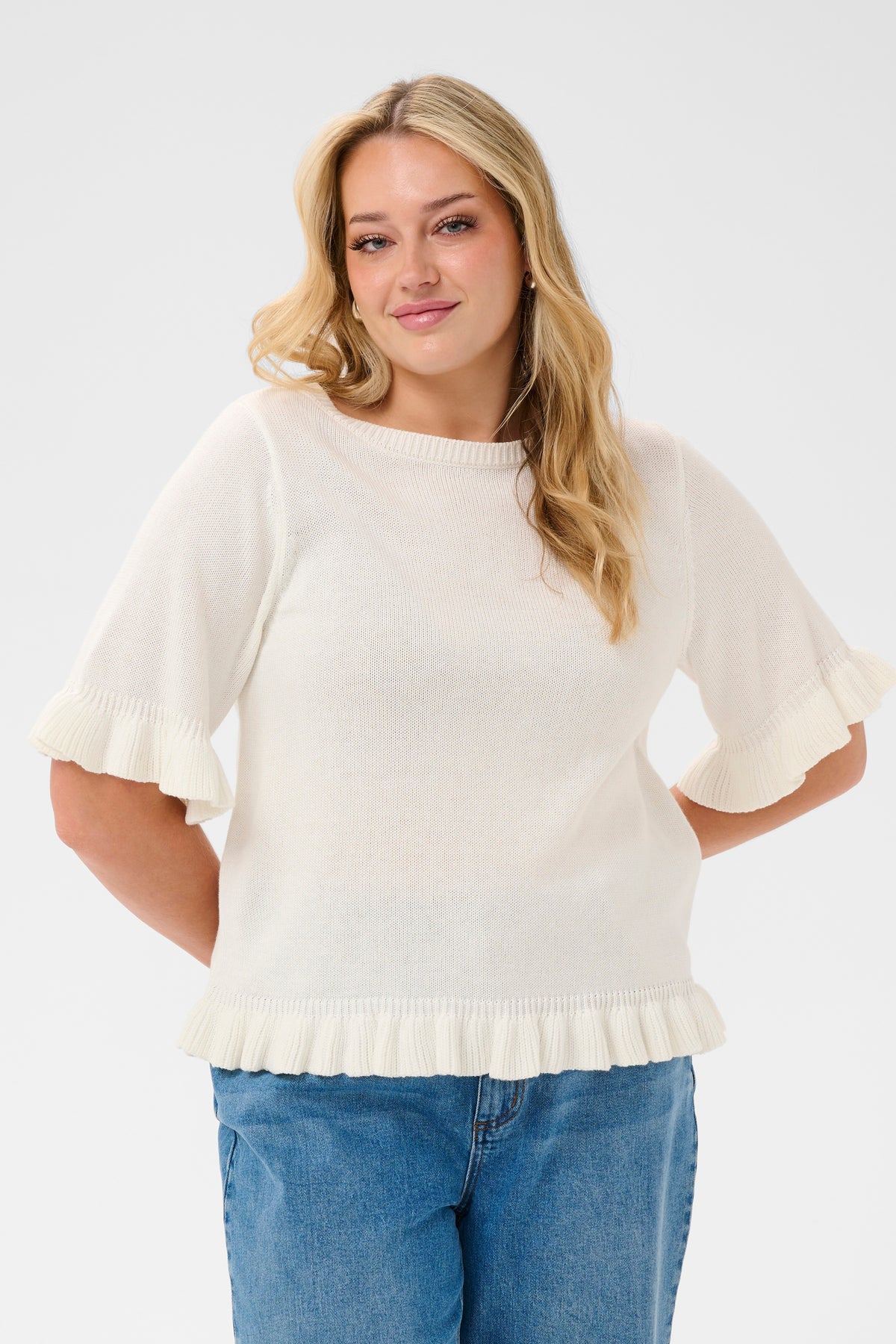 Woman wearing the Kaffe Curve Lindsy Top in White, a white ruffled sweater and blue jeans on a white background