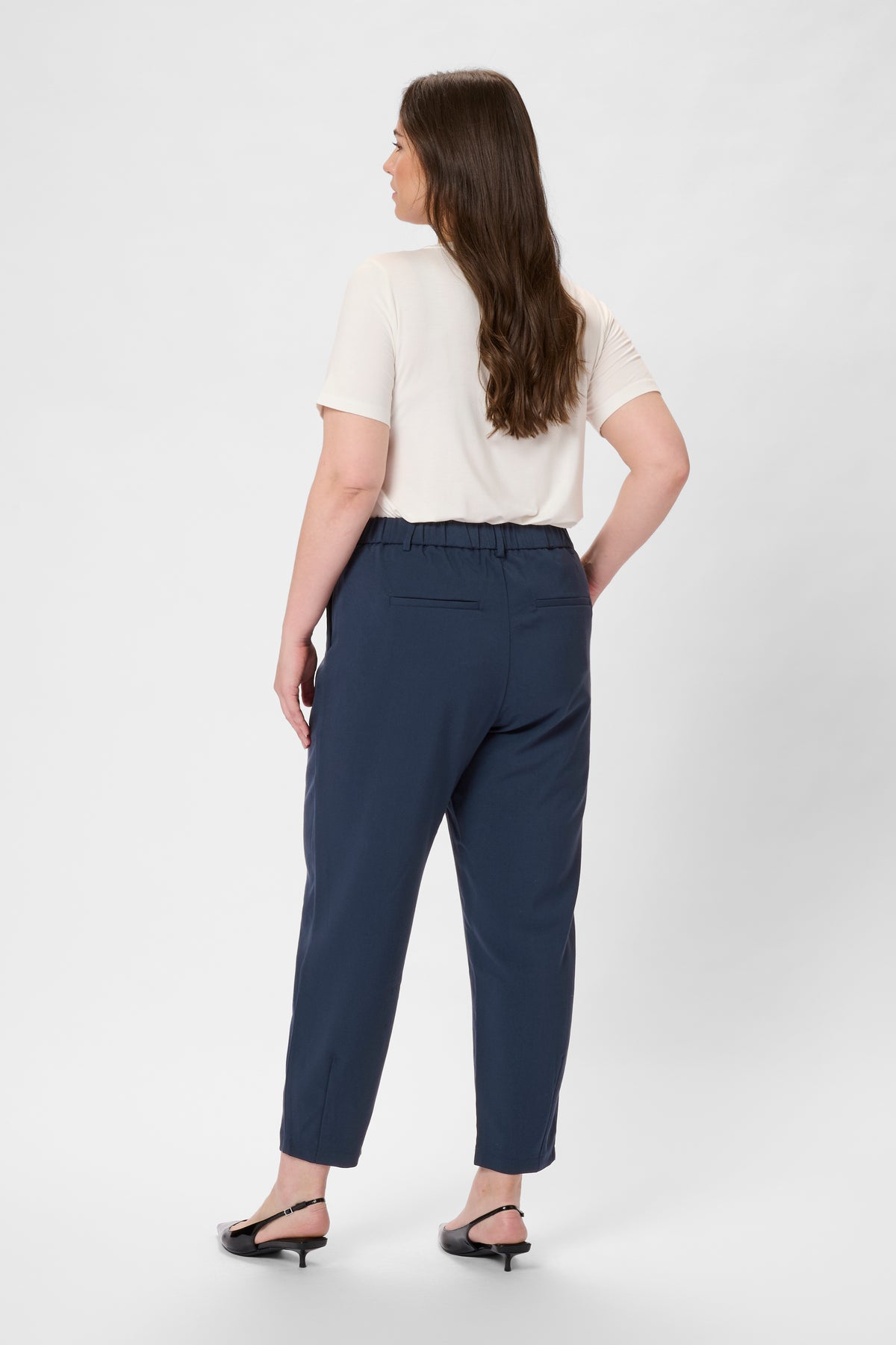 Woman wearing navy ankle length trousers and a white top on a white background