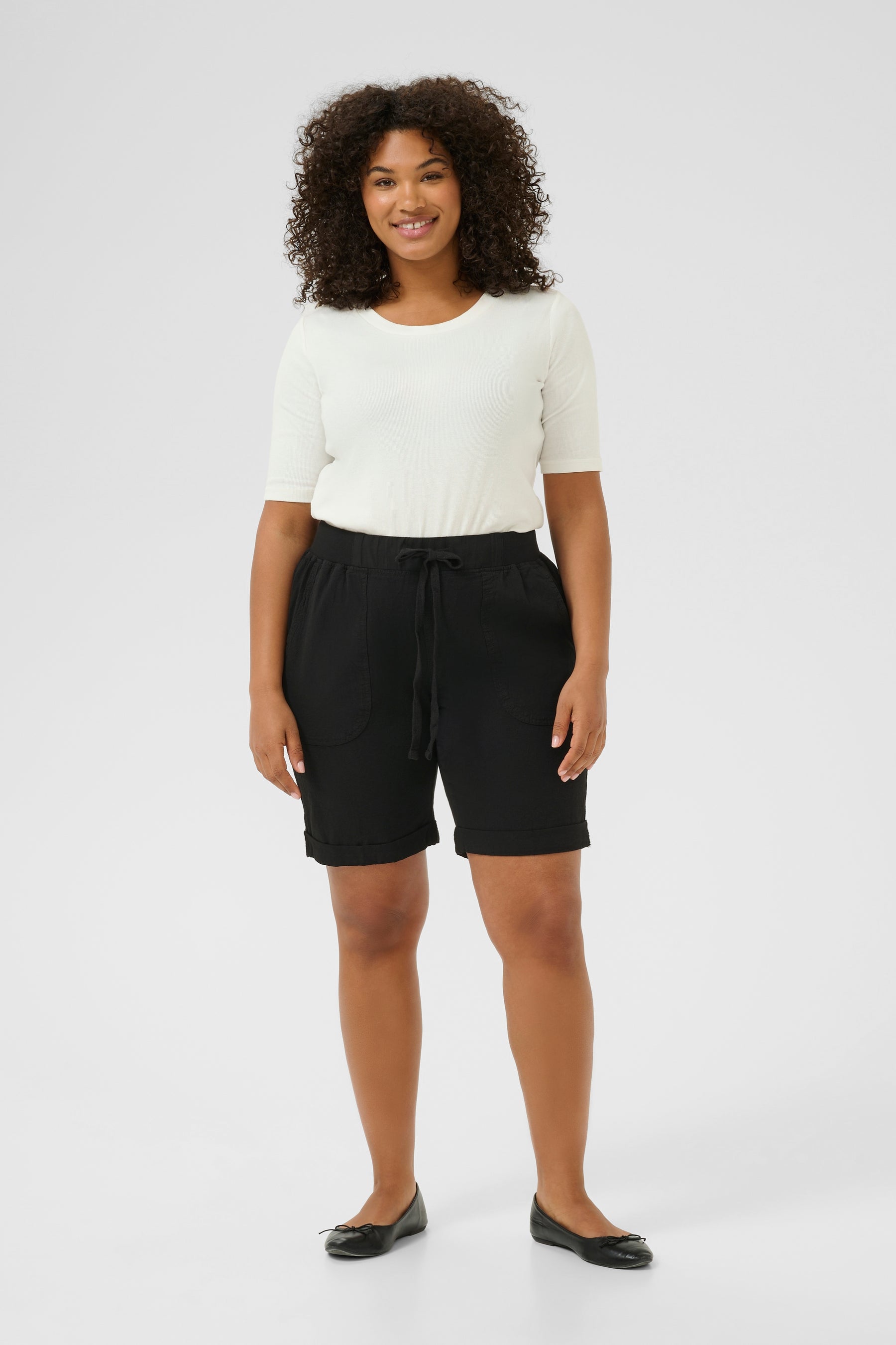 Woman wearing a white top and knee length black shorts with an elasticated waist with drawstrings on a white background