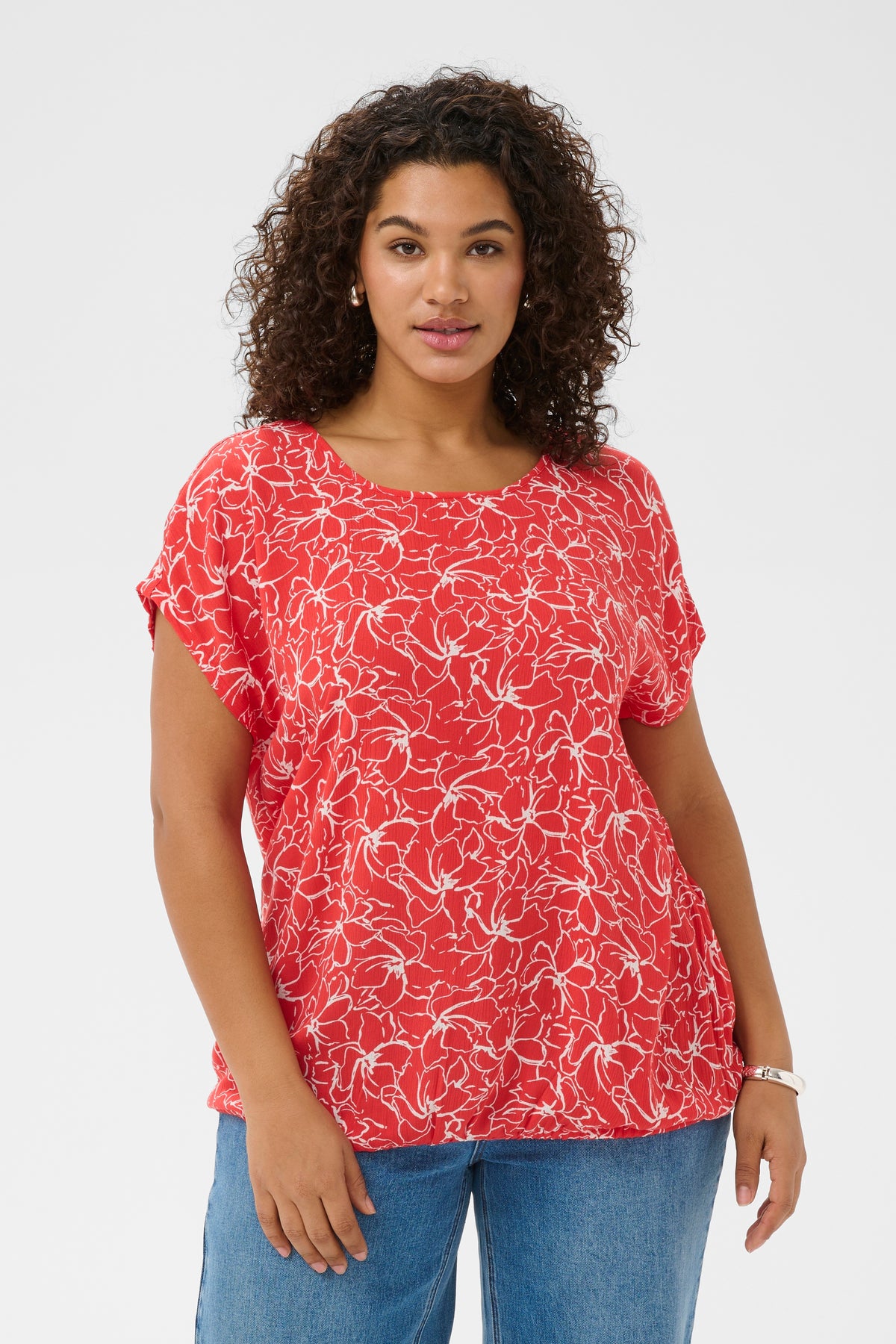 Woman wearing a red floral blouse with a round neckline and short sleeves and blue jeans on a white background