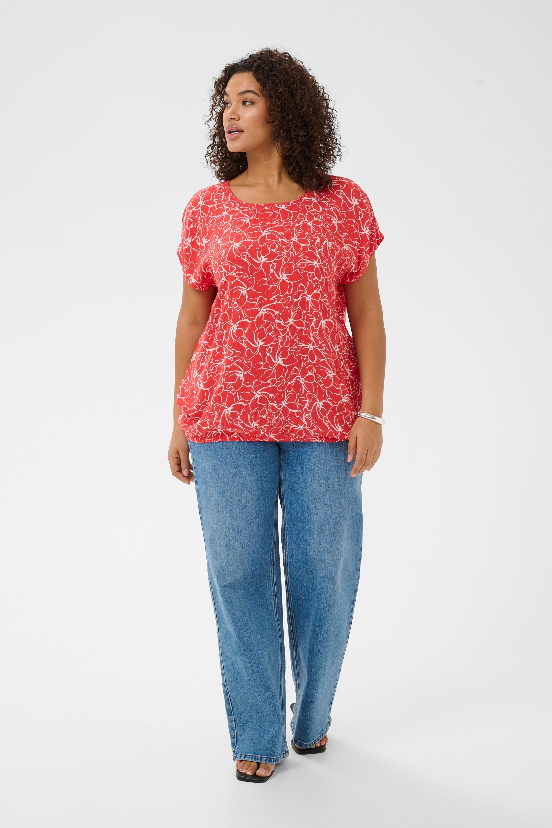 Woman wearing a red top with a white floral design all over, round neckline, short sleeves and elasticated hem and blue jeans on a white background