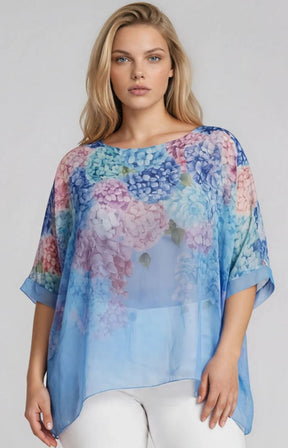 Woman wearing a blue, flowy, floral blouse with a floral design across the top, a round neckline and 1/2 length sleeves against a plain background