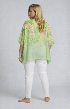 Woman wearing a light green floral blouse with 1/2 length sleeves over a white outfit on a plain background