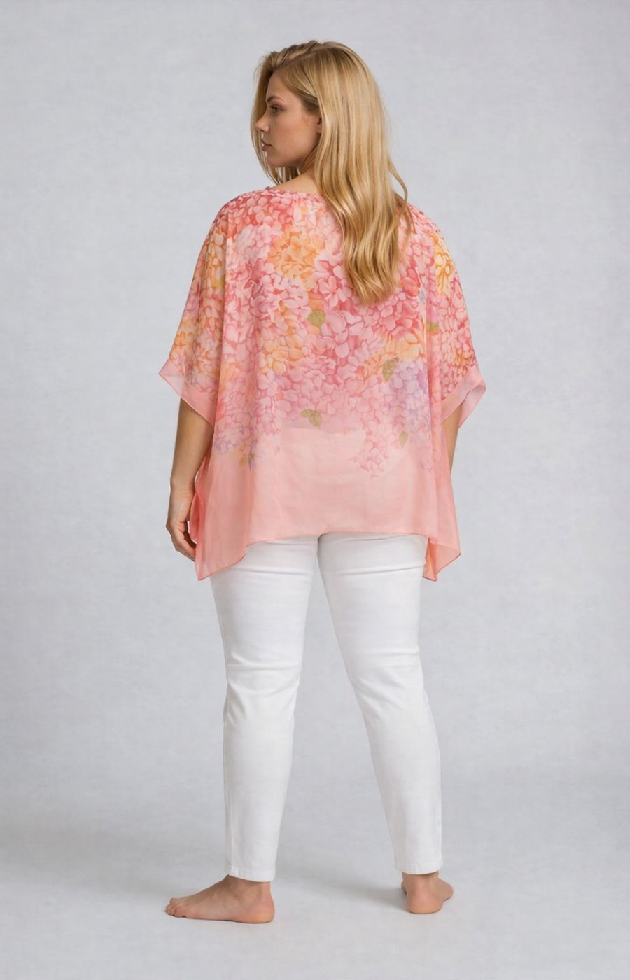 Woman wearing a pink floral, flowy chiffon feel blosure with a floral design and 1/2 length sleeves over white jeans on a plain background
