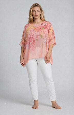 Woman wearing a pink floral blouse with a round neckline and 1/2 length sleeves and white jeans on a plain background