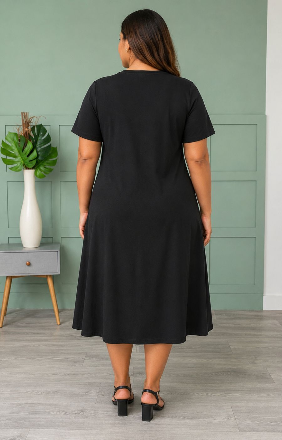 Woman wearing the Leah Dress in Black, a black dress standing in a room with a green wall and a vase on a table.