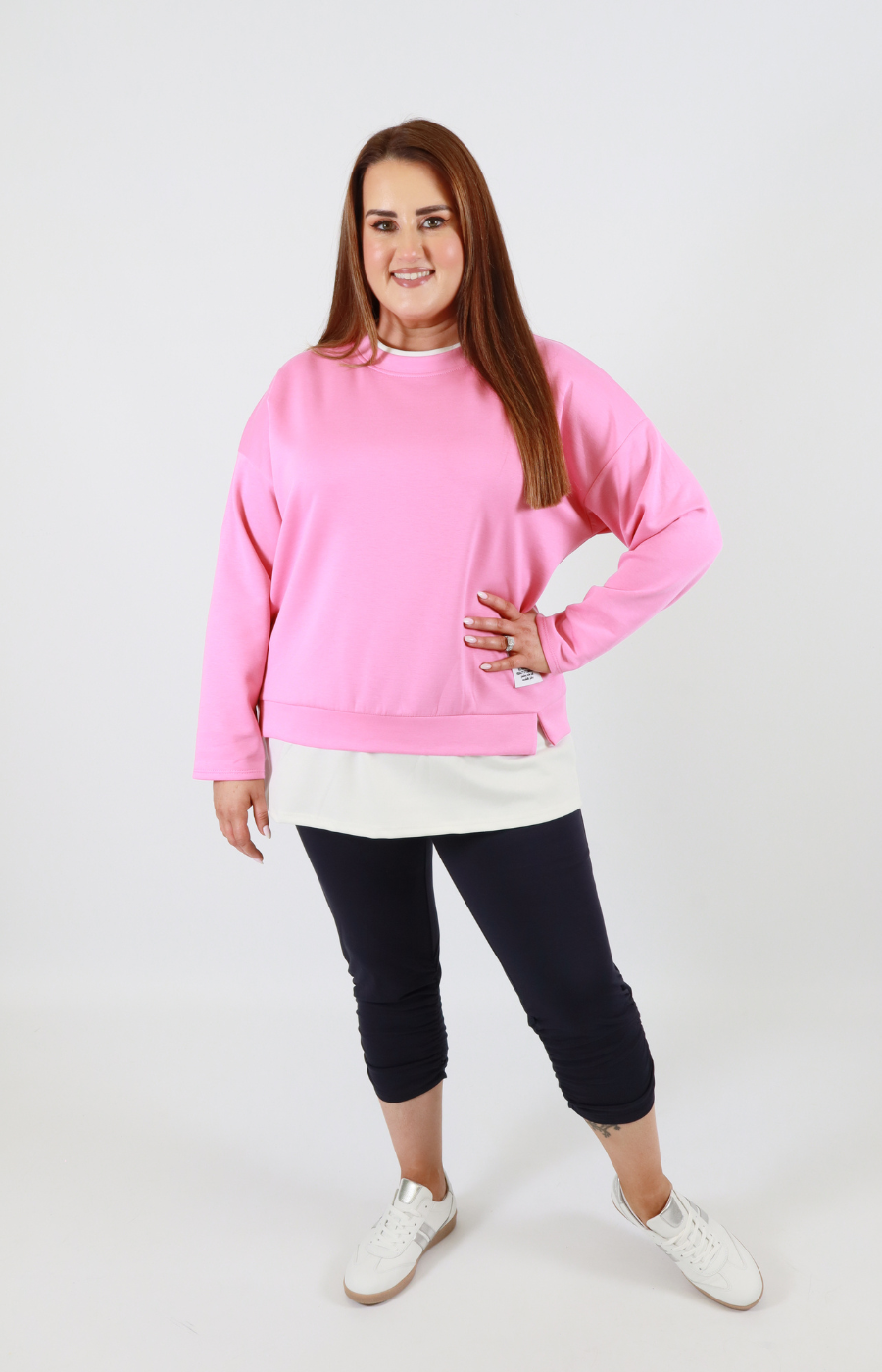 Woman wearing a pink sweatshirt, white shirt, black pants, and white shoes on a white background