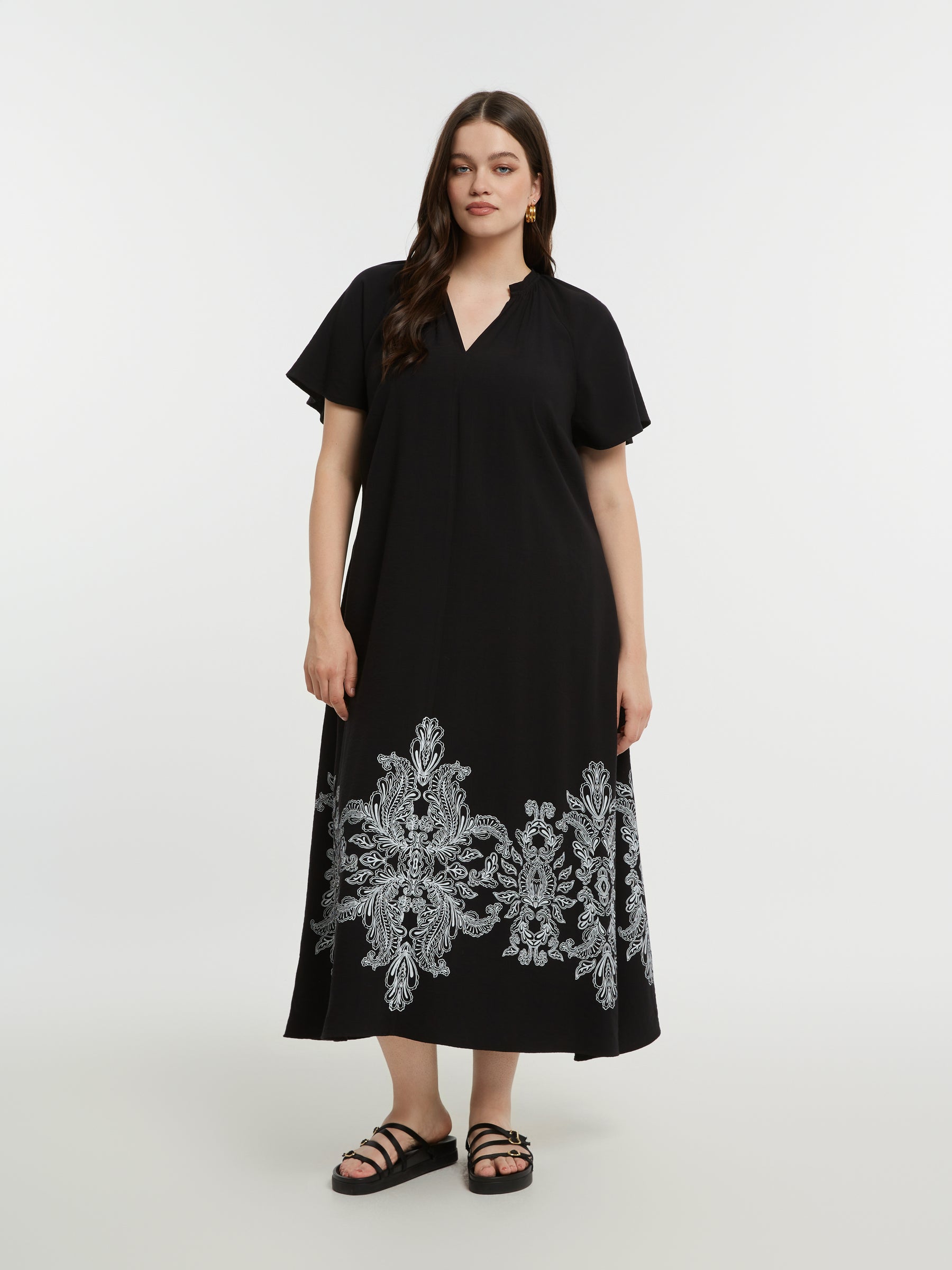 Woman wearing the Mat Black Dress with Paisley Print, a black midi dress with white embroidery, with an A-line shape, V neckline and short sleeves on a white background