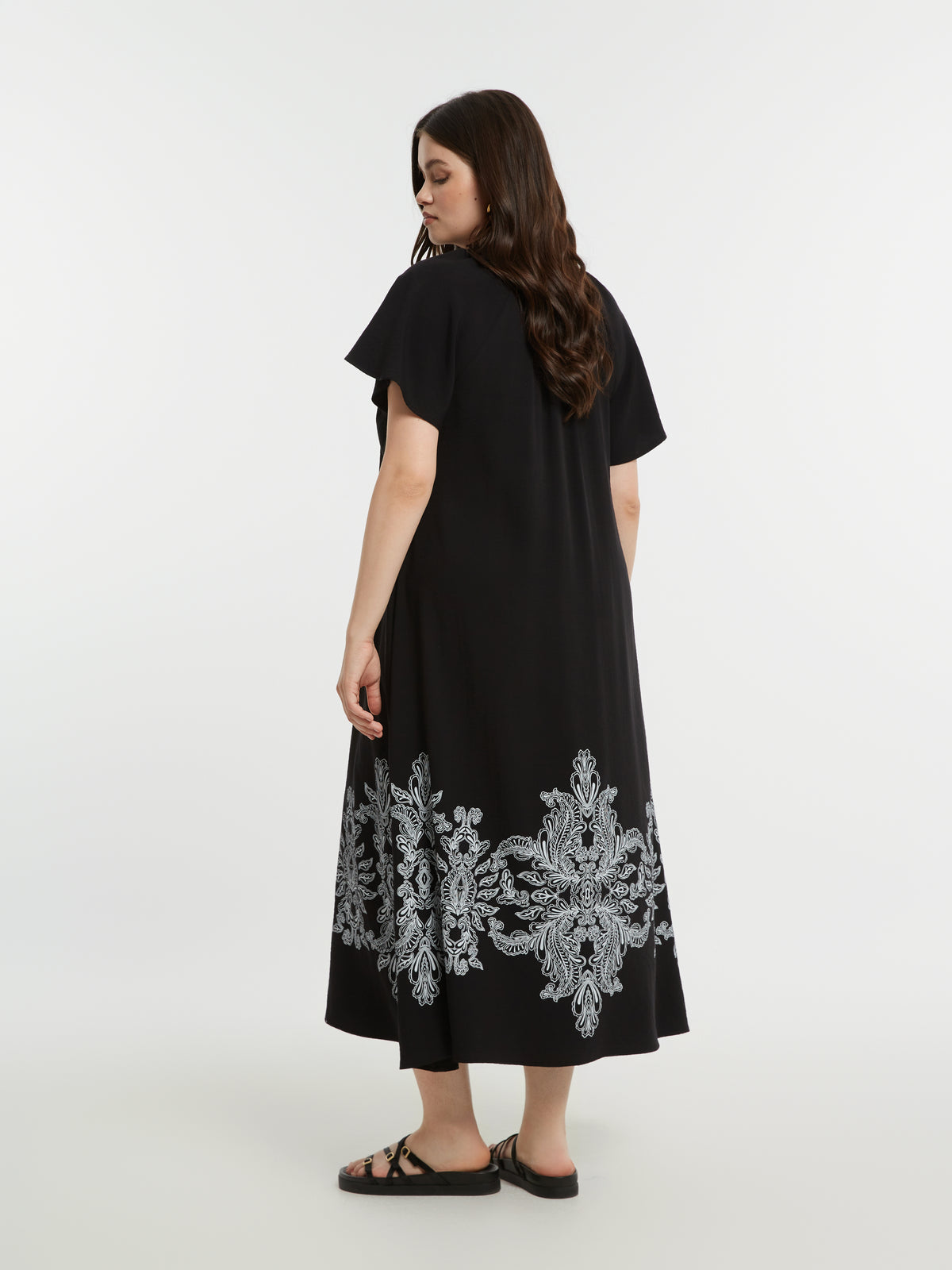 Woman wearing the Mat Black Dress with Paisley Print, a black A-line midi dress with white embroidery at the bottom and short sleeves on a white background