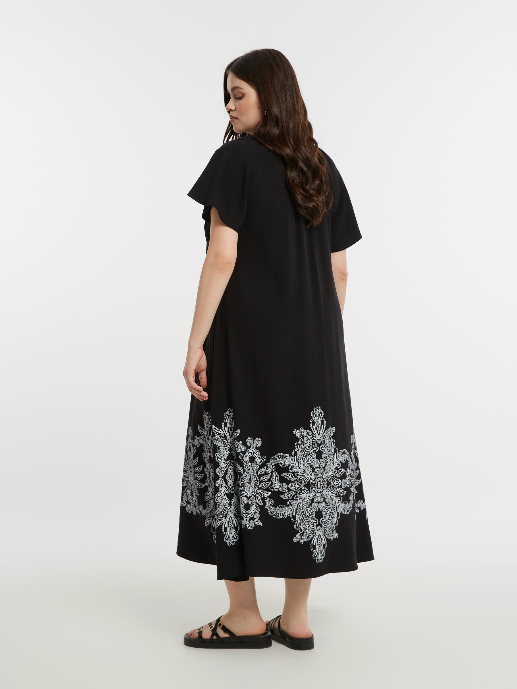 Woman wearing the Mat Black Dress with Paisley Print, a black A-line midi dress with white embroidery at the bottom and short sleeves on a white background