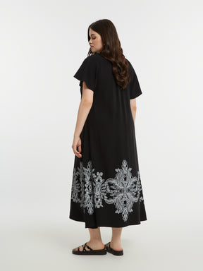 Woman wearing the Mat Black Dress with Paisley Print, a black A-line midi dress with white embroidery at the bottom and short sleeves on a white background