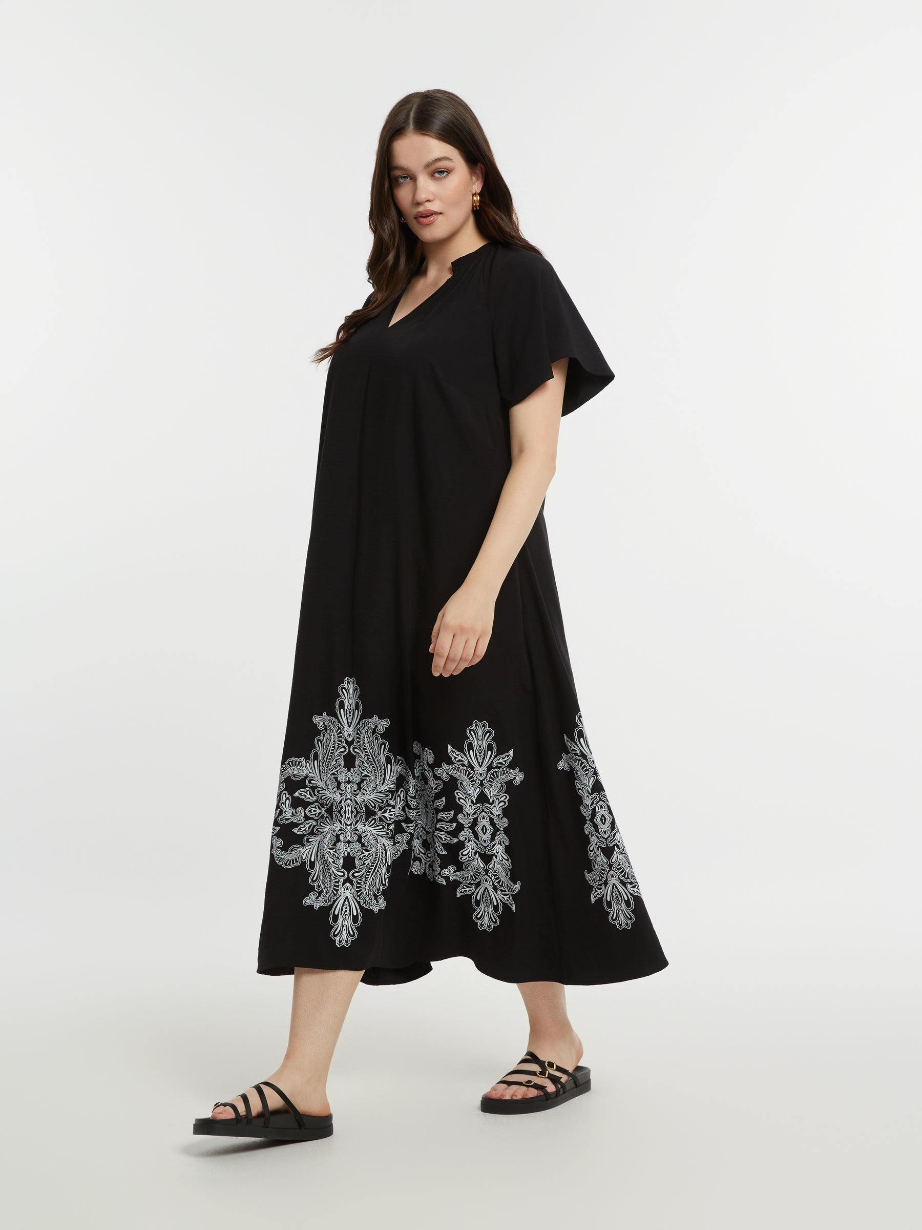 Woman wearing the Mat Black Dress with Paisley Print, a black a-line, midi dress with white embroidery on the bottom, short sleeves and a V neckline on a white background