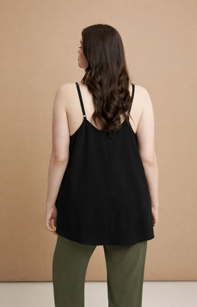 Woman wearing the Mat Cami in Black, a black cami top with spaghetti straps and an A-line shape 
 and green pants against a beige background