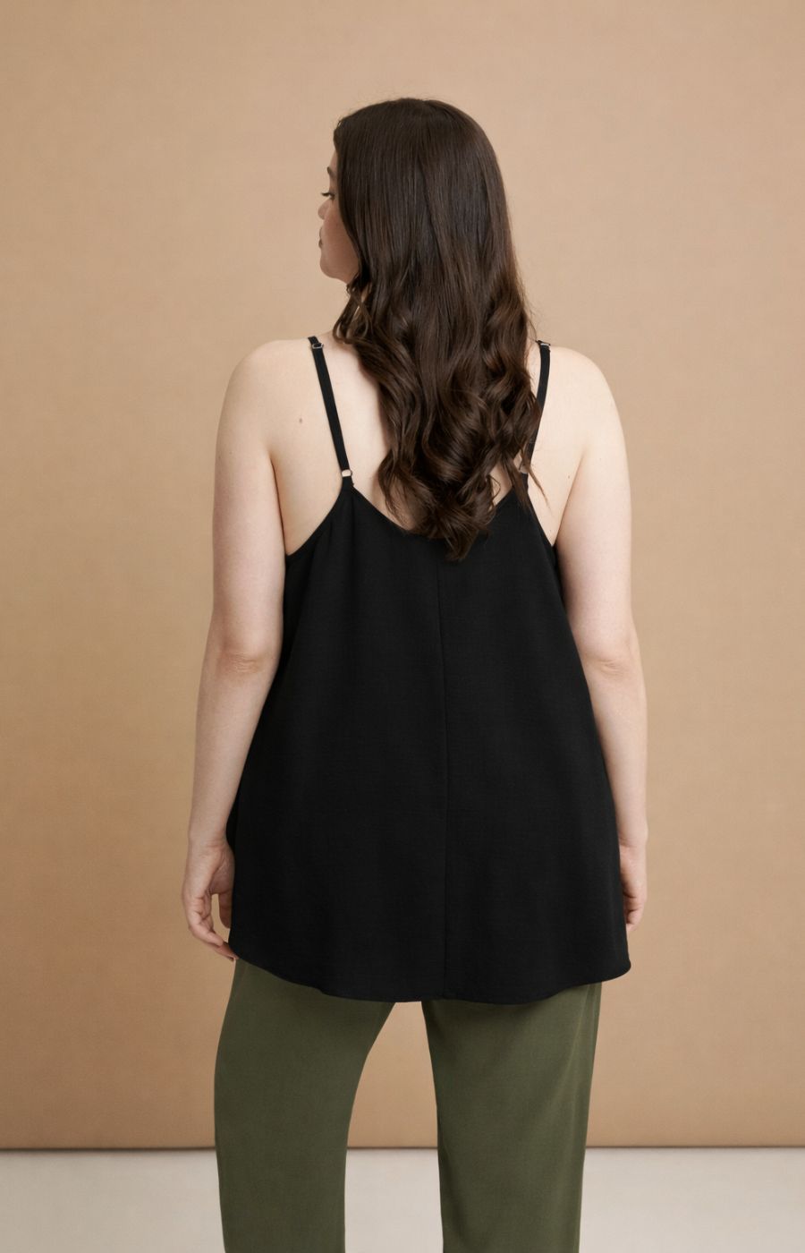 Woman wearing the Mat Cami in Black, a black cami top with spaghetti straps and an A-line shape 
 and green pants against a beige background