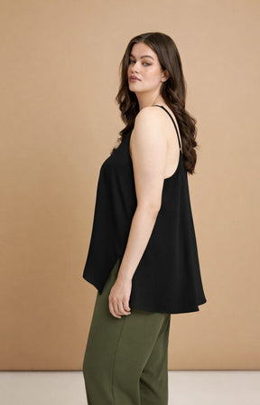 Woman wearing the Mat Cami in Black, a black sleeveless top with an A-line shape and side slits with green trousers
 against a beige background