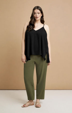 Woman wearing the Mat Cami in Black,  a black cami to with spaghetti straps, a V neckline and a curved hem with side slits and green pants against a beige background