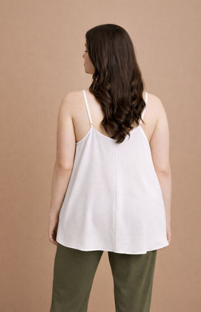 Woman wearing the Mat Cami in White, a white sleeveless top with a V neckline and side slits and green pants against a brown background