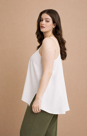 Woman wearing the Mat Cami in White, a white sleeveless top with an Aline shape and side slits  and green pants on a beige background