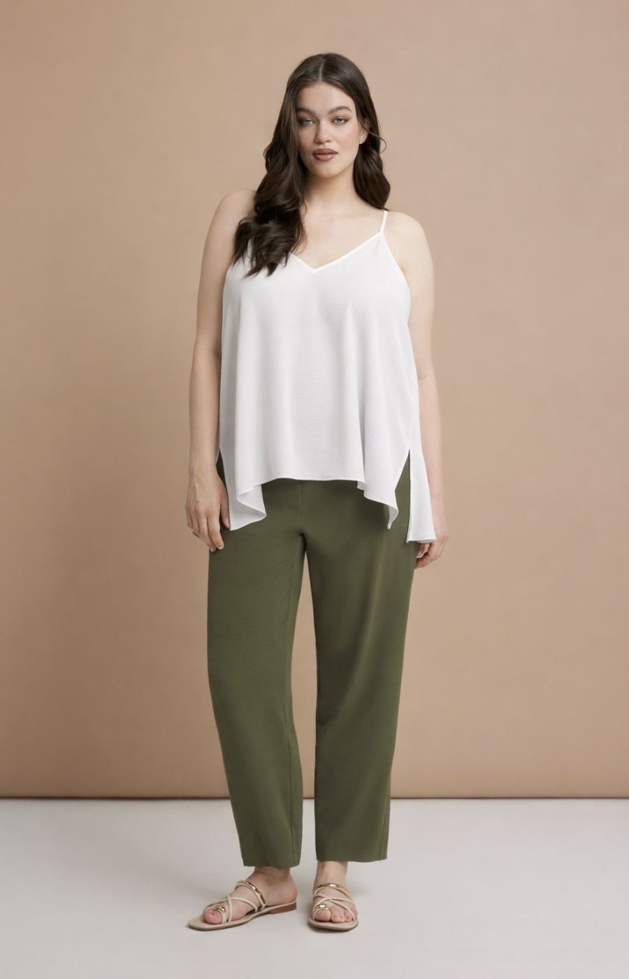 Woman wearing the Mat Cami in White, a white sleeveless top with a V neckline and side slits and green pants against a beige background