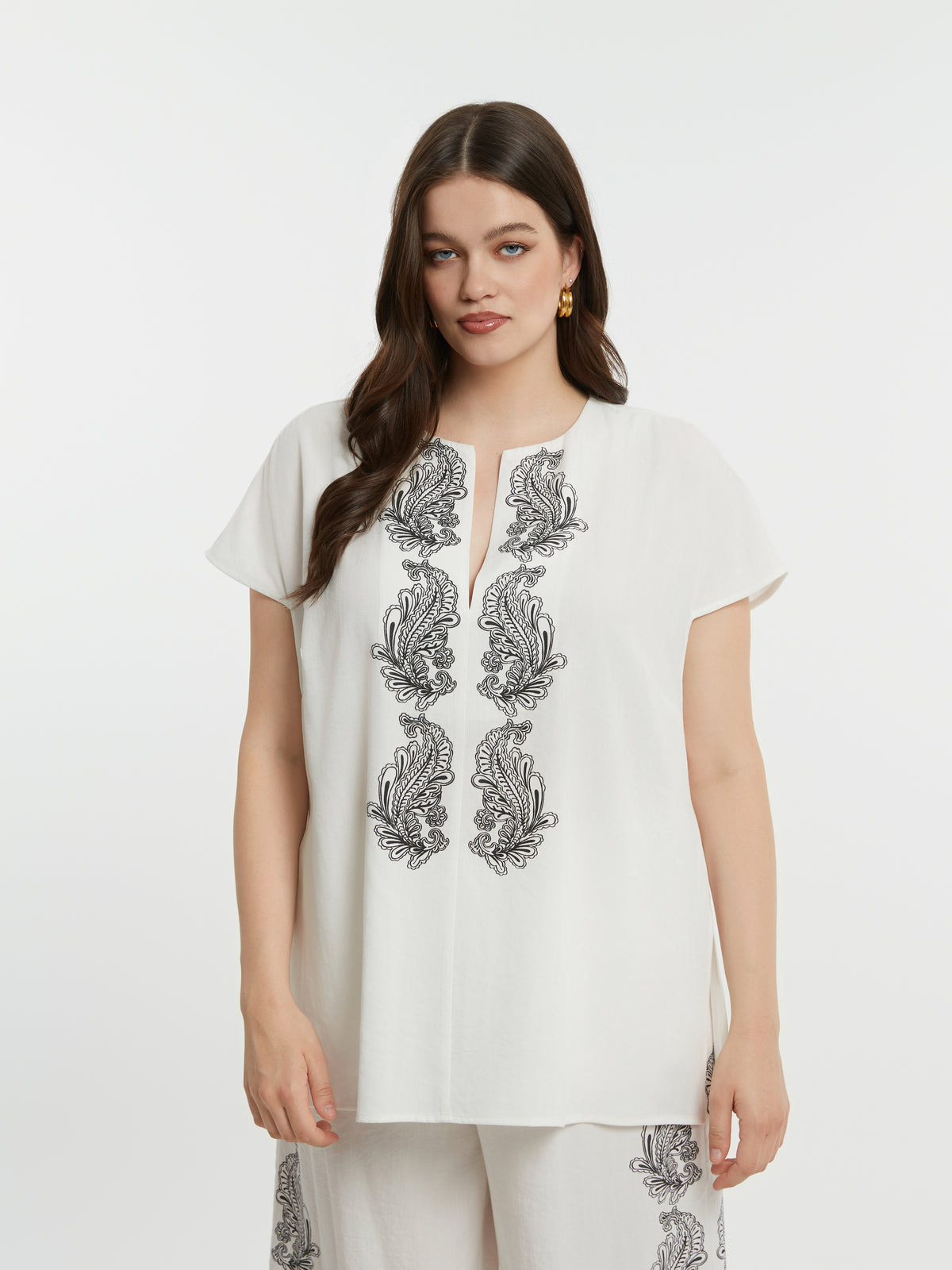 Woman wearing a white top with black floral embroidery with short sleeves and a round neckline with a V slit on a plain background 