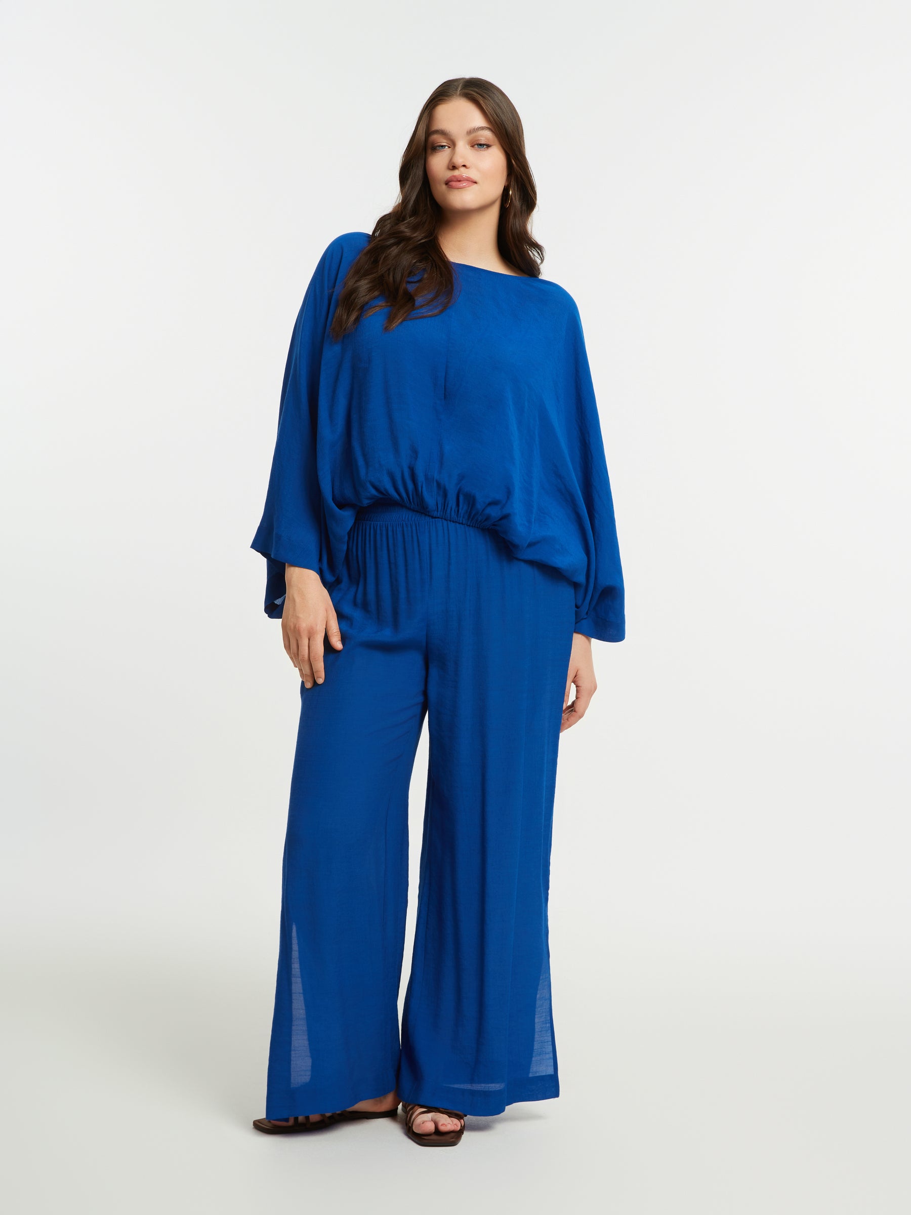 Woman wearing the Mat Slit Trouser in Blue, a high rise trouser with a wide leg and side slits, worn with a blue blouse on a white background