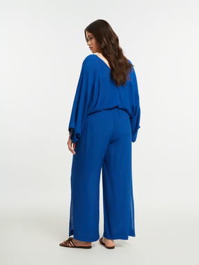 Woman wearing the Mat Slit Trouser in Blue, a blue, wide leg trouser with a pull up style and side slits, worn with a blue blouse on a white background