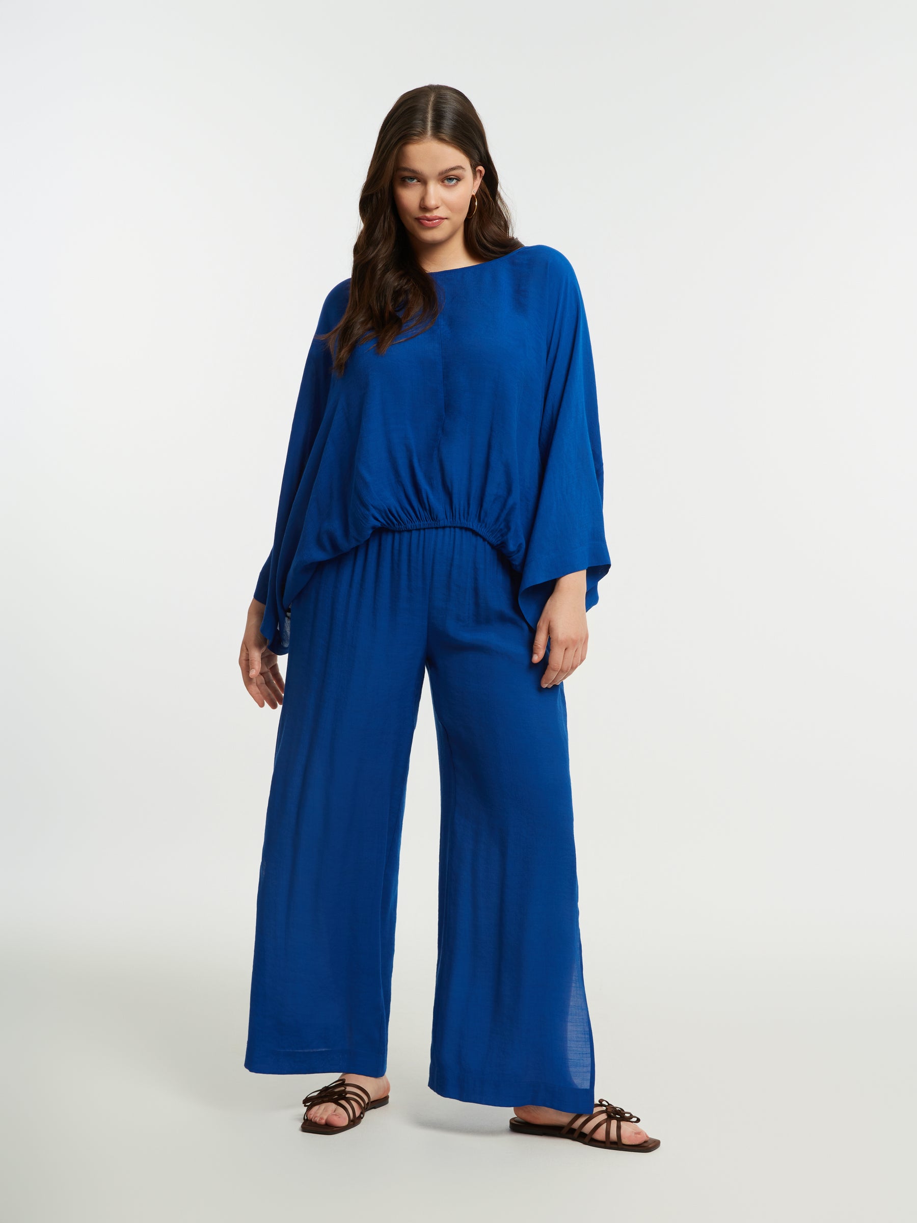 Woman wearing the Mat Slit Trouser in Blue, a wide leg trouser with an elasticated waist and side slits, worn with a blue blouse on a white background
