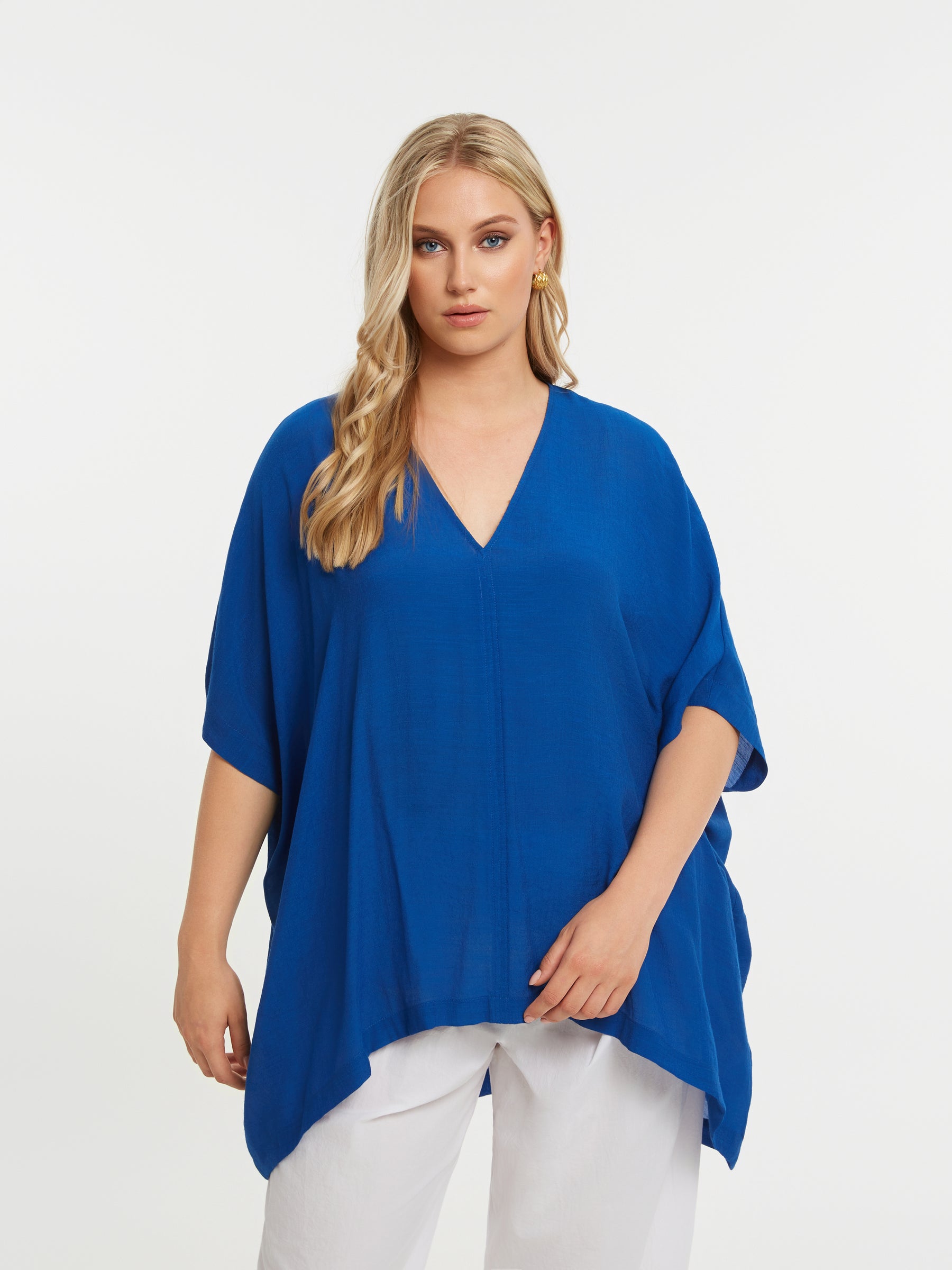 Woman wearing the Mat V Neck Blouse in Blue, a blue top with a V neckline and curved hem, and white pants on a white background