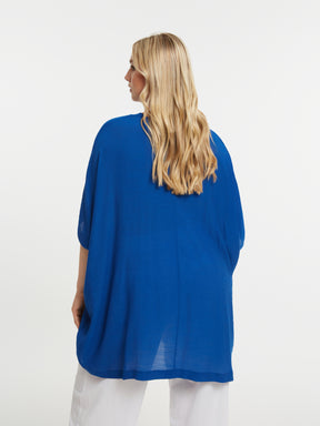 Woman wearing the Mat V Neck Blouse in Blue,  a blue blouse with short sleeves, V neckline and a curved hem, and white trousers on a white background