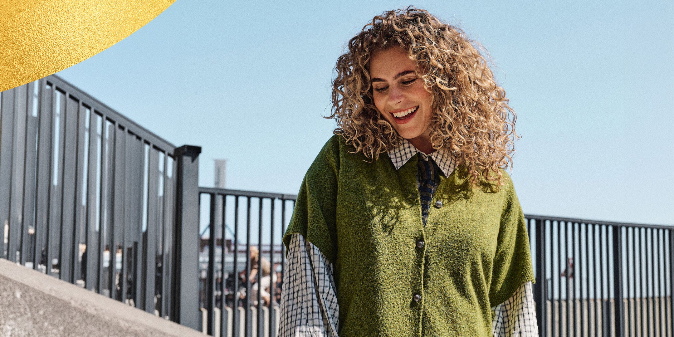 A woman with curly blonde hair smiling outdoors, wearing a green knit poncho-style cardigan over a plaid button-up shirt. She is standing near a black metal railing under a clear blue sky.